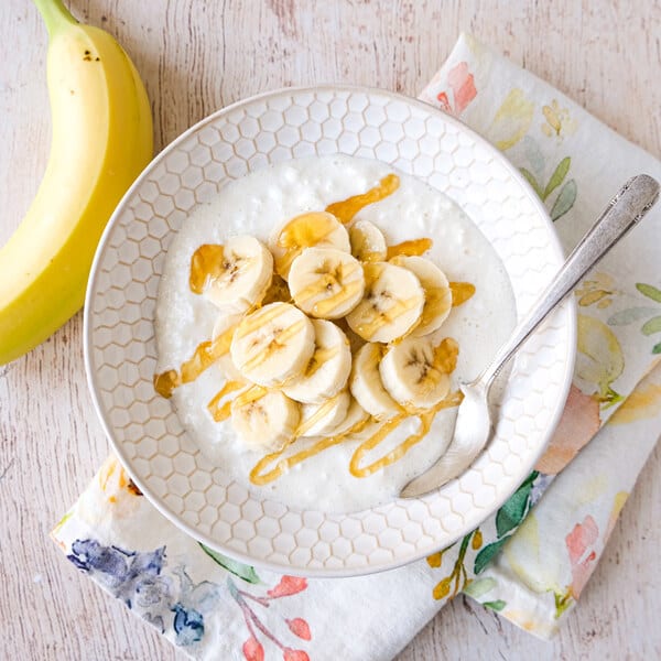Bowl of creamy porridge topped with banana slices and honey drizzle, with a whole banana and floral napkin.