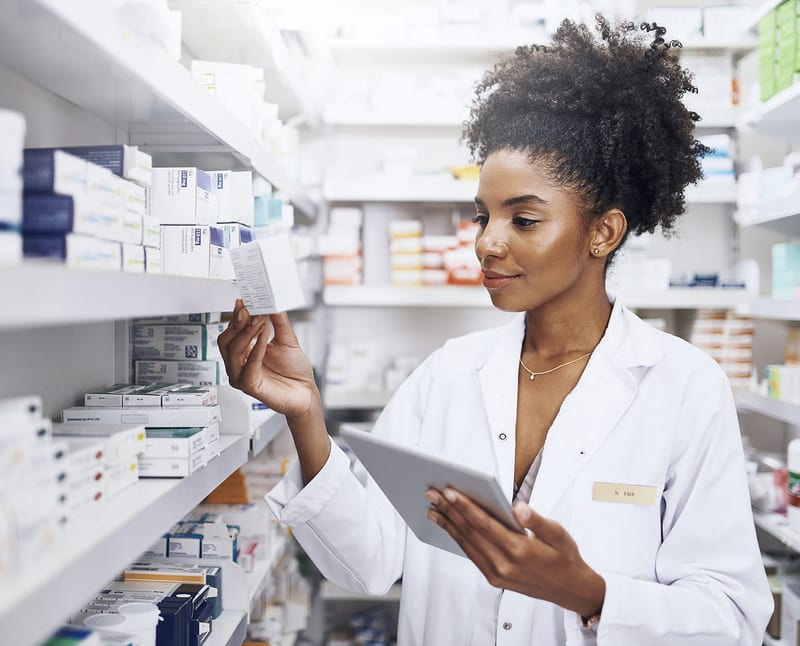 Female pharmacist in white coat holds a tablet and checks medicine box on pharmacy shelf.