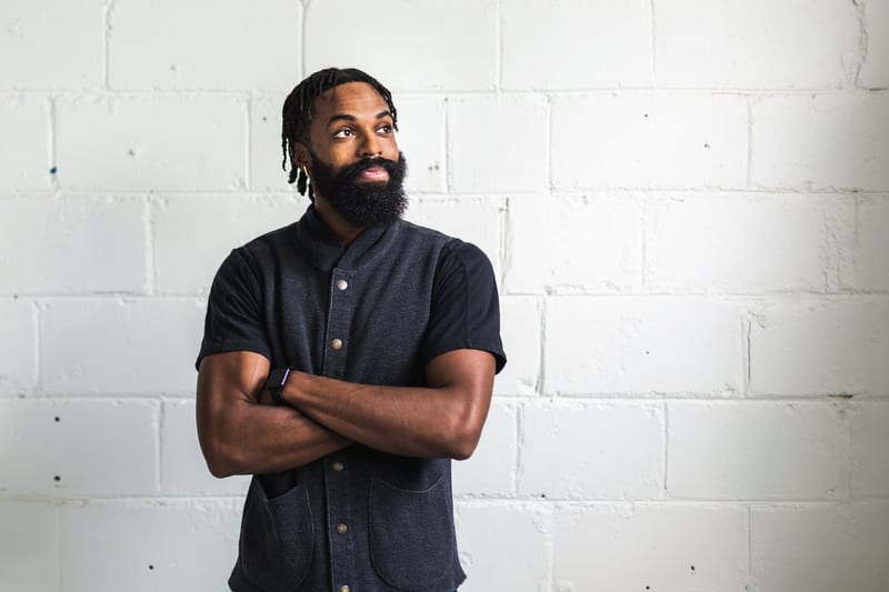a man with a beard is standing in front of a white brick wall with his arms crossed