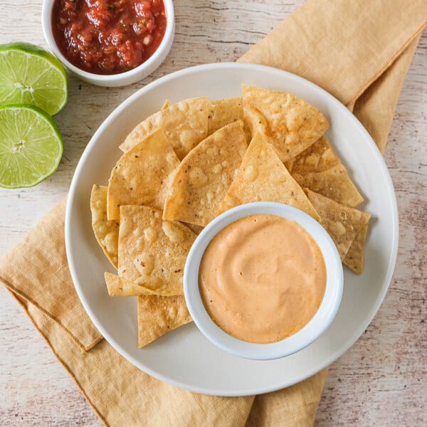 Tortilla chips on a white plate with creamy dip and salsa, lime wedges, and a tan napkin on a rustic table.