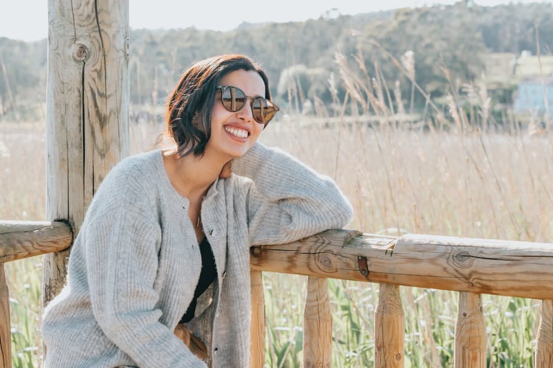 a woman wearing sunglasses and a grey sweater smiles while leaning on a wooden railing
