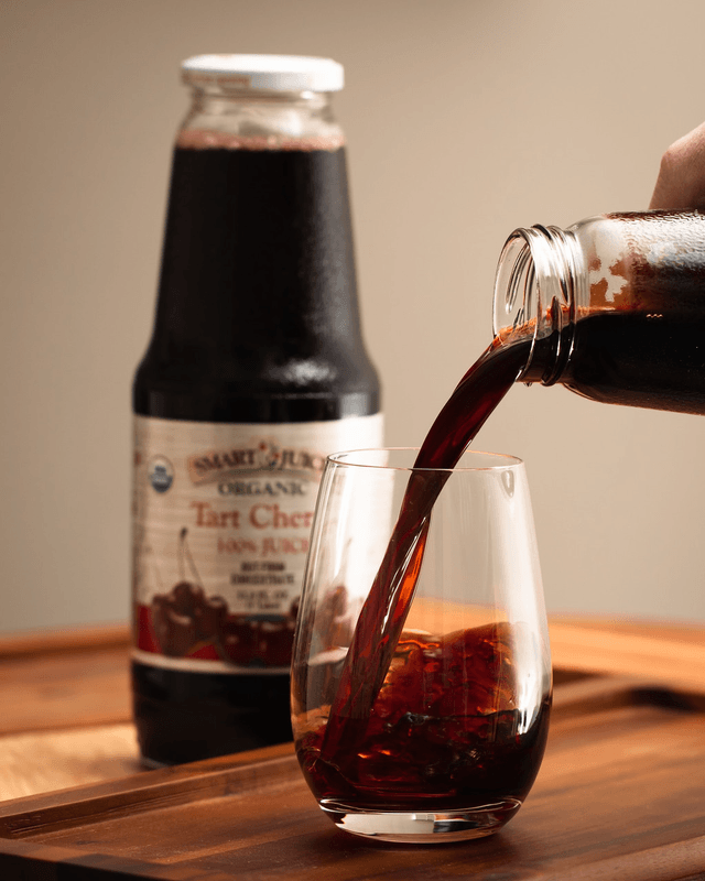 Tart cherry juice poured from bottle into stemless glass on wooden tray, another bottle behind